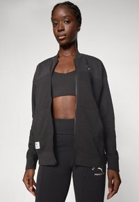 Sort lynlåsjakke i tekstureret stof med ribbede manchetter og sidelommer, båret over en matchende sort sportsbh og leggings.
