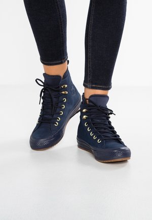 Navy blue high-top sneakers with black laces and gold eyelets worn with dark skinny jeans on a plain white background.
