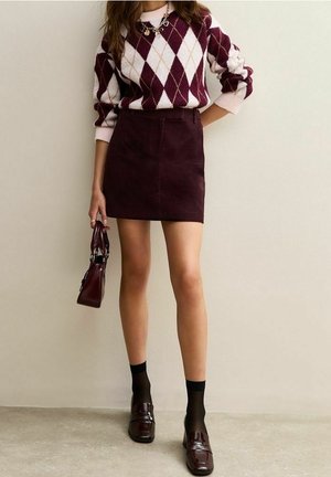 Femme portant un pull à losanges bordeaux et blanc, une mini-jupe bordeaux foncé, des chaussettes noires, des mocassins bordeaux brillants et tenant un petit sac à main bordeaux.