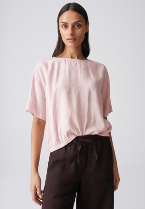 Vrouw met lang donker haar, gekleed in een lichtroze blouse met korte mouwen en zwarte broek met trekkoord, staand tegen een effen achtergrond.