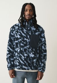 Blauwe en zwarte fleece trui met abstract patroon, hoge kraag, halve rits aan de voorkant en een borstzak in effen zwart. Gestructureerde stof.