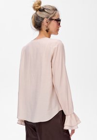 Jasnoróżowa bluzka z długimi rękawami i falbankami przy mankietach, wykonana z gładkiego materiału, ma luźny krój oraz dekolt w łódkę.