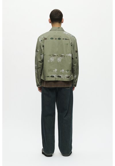Chaqueta de mezclilla verde con un patrón de salpicaduras de pintura, combinada con una camisa marrón oscuro debajo y jeans azules holgados. Diseño minimalista, tejido texturizado.