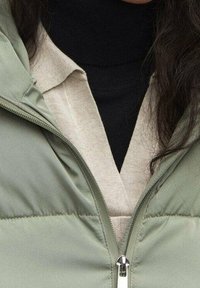 Hellgrüne Steppjacke mit Reißverschluss, getragen über einem schwarzen Rollkragenpullover und einem beigen Hemd, zeichnet sich durch gesteppte Textur und Nahtdetails aus.