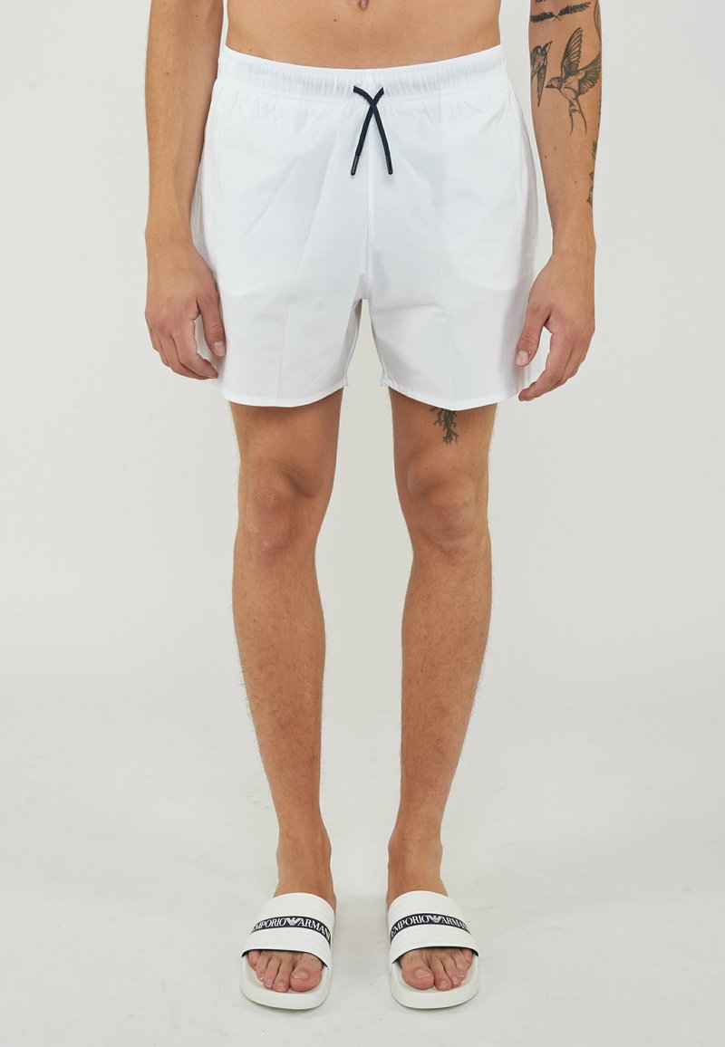 Homme portant un short de bain blanc avec un cordon noir et des sandales slides blanches avec des brides noires avec logo, montrant des tatouages sur les bras et la jambe.