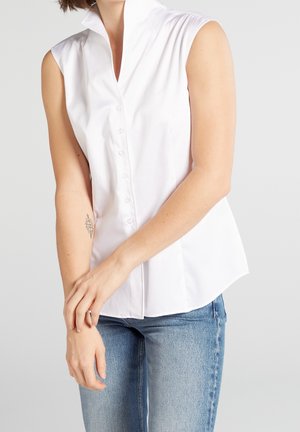 Mouwloze witte blouse met knopen gecombineerd met lichtblauwe spijkerbroek, model met tatoeage op linker onderarm, armen losjes gekruist voor het lichaam.