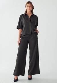 Camicia nera, ampia e comoda, con maniche corte arrotolate, colletto a punta e chiusura con bottoni. Abbinata a pantaloni neri a gamba larga e tacchi.