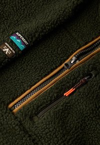 Dunkelgrüne Fleecejacke mit einer großen Zipper-Tasche mit orangefarbenen Akzenten und einem Superdry-Logopatch. Weiche Textur und strapazierfähiges Design.