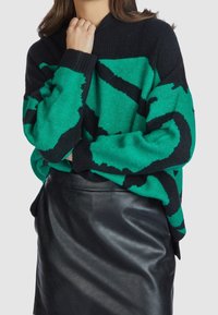 Kobieta w zielonym i czarnym swetrze w wzory, zestawionym z czarną skórzaną spódnicą, pozująca z jedną ręką blisko podbródka.