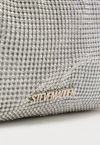 Strieborná večerná kabelka pokrytá kamienkami, s metalickým zlatým logom "Steve Madden" a hladkou textúrou. Kompaktný obdĺžnikový tvar.