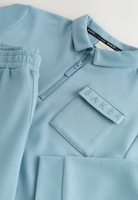 Himmelblaues Sportoberteil mit gerippter Textur, Kragen, Reißverschluss und einer aufgenähten Tasche mit der Aufschrift "BAKER."