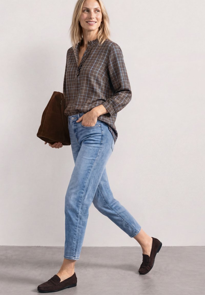 Femme marchant portant une chemise à carreaux marron, un jean bleu clair, des mocassins marron foncé et tenant un grand sac marron.