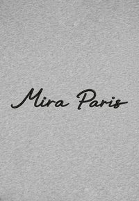Broderie noire en cursive indiquant "Mira Paris" sur un fond de tissu gris clair.
