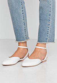 Białe skórzane baleriny z wąskim noskiem, wycięciem oraz paskiem wokół kostki. Gładka faktura z płaską podeszwą, w połączeniu z dżinsami z postrzępionym dołem.