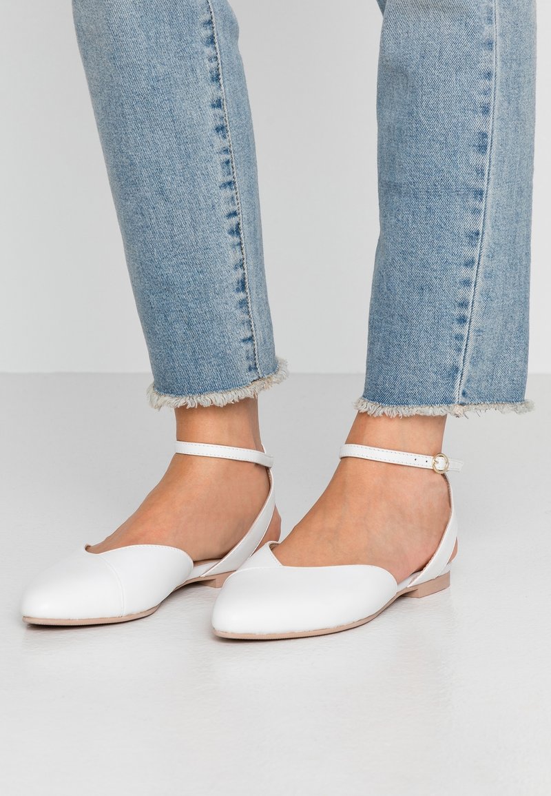 Białe skórzane baleriny z wąskim noskiem, wycięciem oraz paskiem wokół kostki. Gładka faktura z płaską podeszwą, w połączeniu z dżinsami z postrzępionym dołem.