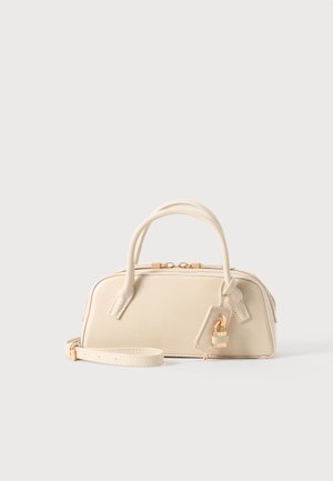 Kleine beige handtas met dubbele handvaten, gouden rits en afneembare band, met een gouden hangslot bedel op een effen achtergrond.