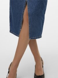 Picioare încrucișate, purtând o fustă albastru denim cu o despicătură centrală și pantofi cu barete negri, pe un fundal alb.