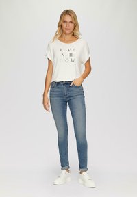 Biely krátky rukáv tričko s textom "ŽIJ V PRÍTOMNOSTI", skombinované s modrými priliehavými džínami a bielymi teniskami, s hladkou textúrou.