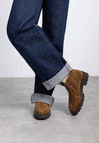 Botines de ante marrón con cordones, que presentan una suela negra gruesa. Llevados con jeans de mezclilla azul oscuro, con los puños doblados para revelar el forro gris.