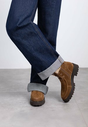 Botines de ante marrón con cordones, que presentan una suela negra gruesa. Llevados con jeans de mezclilla azul oscuro, con los puños doblados para revelar el forro gris.