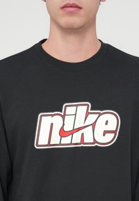 Čierna mikina s okrúhlym výstrihom s veľkým bielym a červeným logom Nike na hrudi, zdôrazňujúcim výrazný text a dizajn.