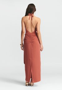 Rudai spalvos maksi suknelė su kaklo juosta, atvira nugara, raukšlėmis juosmens srityje ir šoniniu įpjovimu. Švelni medžiaga su minkšta tekstūra.
