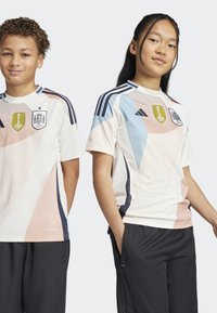 Twee personen dragen een wit voetbalshirt met pastel geometrische vormen, contrasterende marineblauwe accenten, en de FIFA- en Spanje-emblemen.