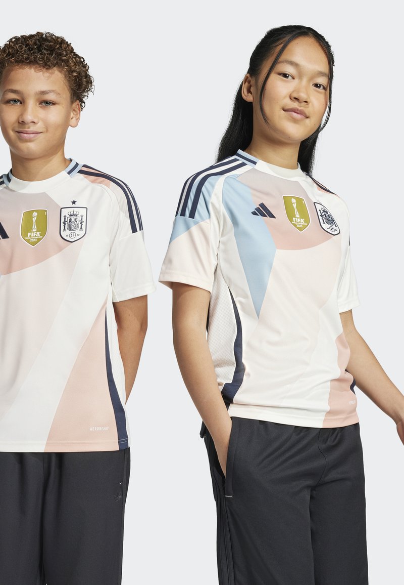 Twee personen dragen een wit voetbalshirt met pastel geometrische vormen, contrasterende marineblauwe accenten, en de FIFA- en Spanje-emblemen.