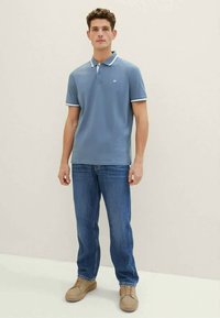 Lyseblå polo med hvide detaljer og krave, kombineret med blå jeans og beige sko. Modellen står foran en neutral baggrund.