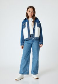 Chaqueta de mezclilla con paneles de forro polar blanco, bolsillos frontales, jeans de pierna ancha color azul claro y zapatillas blancas. En un fondo blanco.