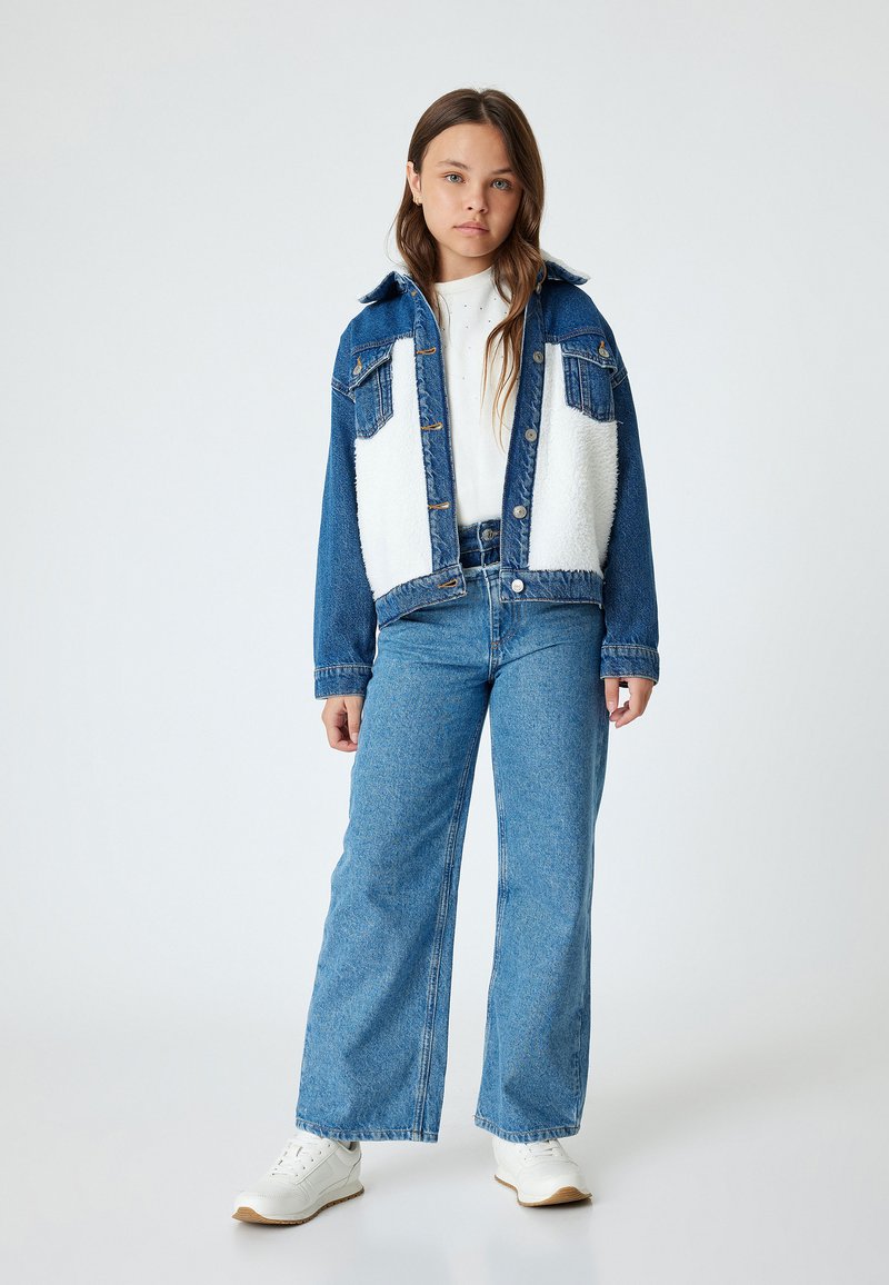 Chaqueta de mezclilla con paneles de forro polar blanco, bolsillos frontales, jeans de pierna ancha color azul claro y zapatillas blancas. En un fondo blanco.