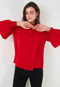 Red blouse with a structured collar, button-down front, and voluminous, gathered sleeves. Smooth fabric texture and relaxed fit.