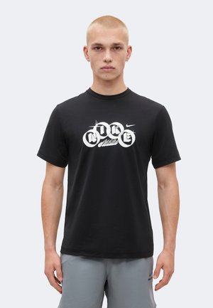 Ung mand med kort blondt hår iført en sort grafisk Nike t-shirt og grå shorts, stående mod en lys ensfarvet baggrund.