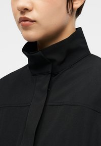 Κοντινό πλάνο ενός ατόμου που φοράει μαύρο μπουφάν με ψηλό γιακά και κουμπιά μπροστά, δείχνοντας τα χείλη, το πηγούνι, τον λαιμό και μερικό αυτί.