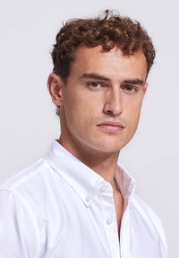 Jonge man met krullend bruin haar, gekleed in een wit overhemd met knopen, die iets opzij kijkt met een neutrale uitdrukking.