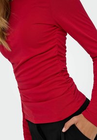 Rode longsleeve top met geraffineerde zijnaad, gemaakt van een zachte stof, met een aansluitende pasvorm en een ronde halslijn. Zwarte onderkleding deels zichtbaar.