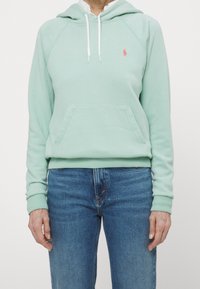Light green hoodie in soft cotton. Features a front pocket, ribbed cuffs, and a white drawstring. Small pink logo on the left chest.
