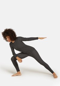 Czarny bodysuit z elastycznego materiału, z białym wzorem falistym, długimi rękawami i dopasowanym krojem. Modelka pozowała w atletycznej pozie, na boso.