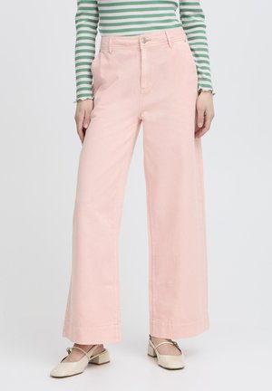 Modella che indossa pantaloni rosa a vita alta e gamba larga con tasche laterali, abbinati a scarpe color crema con tacco basso e una maglia a maniche lunghe a righe verdi e bianche.