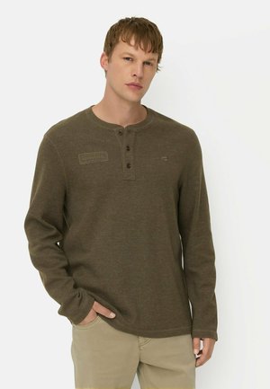 Ung mand iført en olivengrøn langærmet henley-shirt med knapper og beige bukser, stående mod en ensfarvet baggrund.