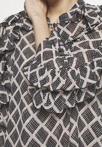 Blusa leggera con motivo geometrico in bianco e nero, caratterizzata da maniche a volant e un dettaglio sul collo a forma di chiave. Realizzata in tessuto trasparente.