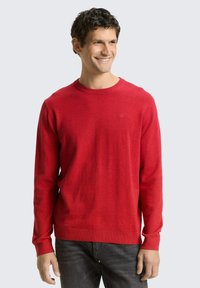 Maglione rosso in maglia con scollo rotondo e maniche lunghe. Tessuto liscio e polsini a coste. Abbinato a jeans grigio scuro.