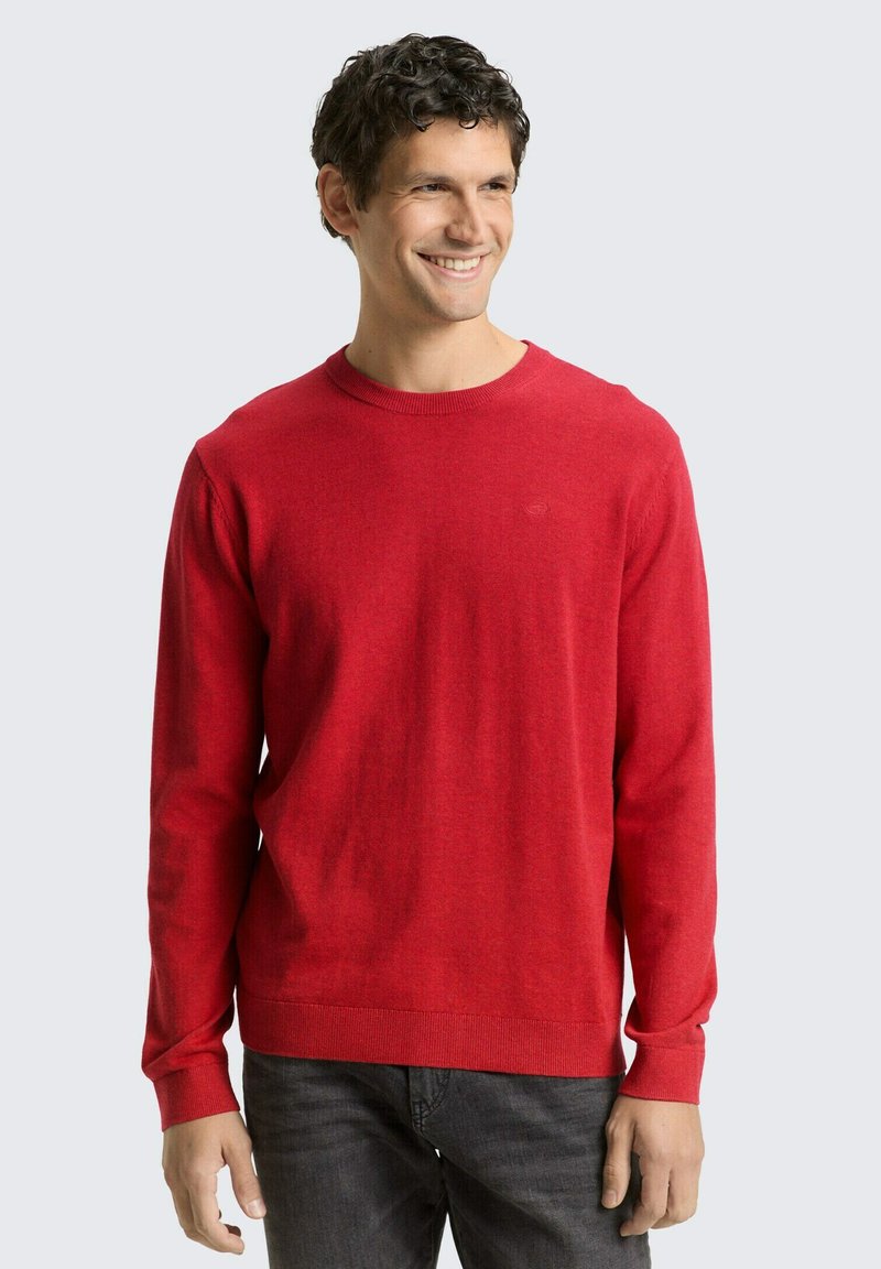 Maglione rosso in maglia con scollo rotondo e maniche lunghe. Tessuto liscio e polsini a coste. Abbinato a jeans grigio scuro.