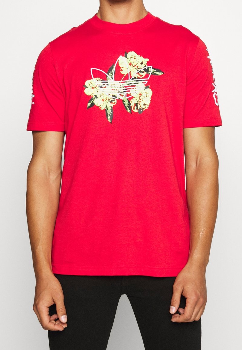 Červená košile s krátkým rukávem a bílým květinovým designem loga Adidas na hrudi, kterou nosí stojící osoba se rukama u těla.