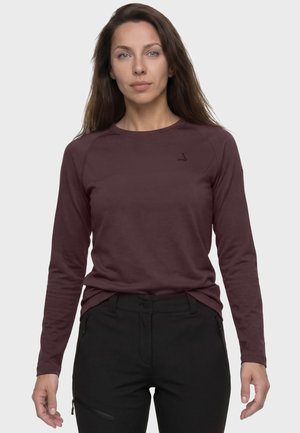 Frau mit langen braunen Haaren, die ein bordeauxrotes Langarmshirt und schwarze Hosen trägt, steht vor einem einfarbigen hellen Hintergrund.