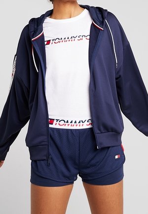 Persoon draagt een marineblauwe zip-up hoodie, een wit Tommy Sport T-shirt en marineblauwe mesh Tommy Sport shorts, staand met de armen licht uitgestrekt.