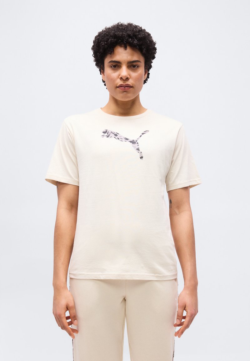 Άτομο με κοντά σγουρά μαλλιά που φοράει ανοιχτό μπεζ μπλουζάκι με σκούρο μοτίβο λογότυπο Puma στο κέντρο του στήθους.