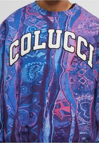 Bunter Pullover mit wirbelnden blauen und lila Mustern. Das "COLUCCI"-Logo ist in weißer Schrift prominent auf der Vorderseite abgebildet.