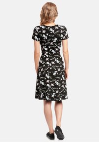 Kobieta z falującymi blond włosami, ubrana w czarną sukienkę w kwiaty z krótkimi rękawami, stoi tyłem do widza, mając na sobie czarne buty.