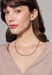 Naszyjniki w odcieniu złota nałożone na siebie; jeden z okrągłymi czarnymi koralikami, a drugi to cienki złoty łańcuszek. Noszone na bluzce z dekoltem w serek.